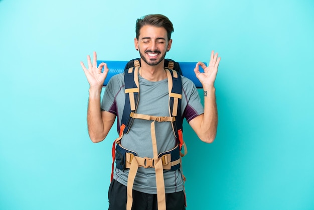 Hombre joven montañero con una mochila grande aislado sobre fondo azul en pose zen