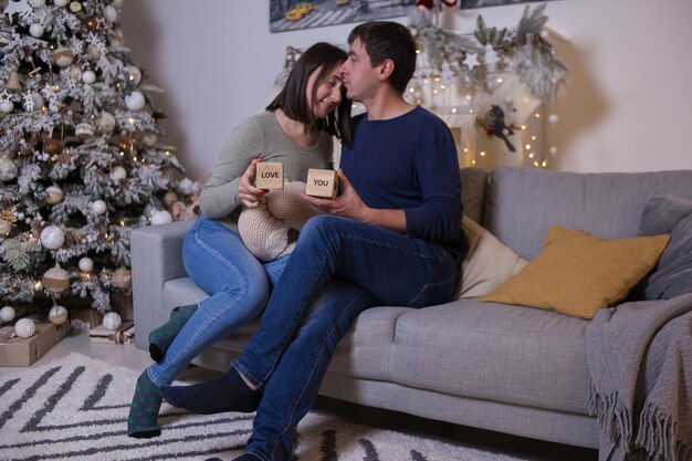 Hombre joven y mujer sentada en el sofá y cerca del árbol de Navidad con regalos y abrazos. Feliz navidad y próspero año nuevo. Amorosa pareja feliz con regalos en el interior de la habitación decorada. Amor. Hogar.