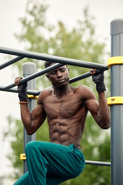 Hombre joven musculoso haciendo ejercicio en el campo deportivo Hombre africano mirando hacia un lado mientras hace ejercicios de barra horizontal Modelo masculino sin camisa haciendo ejercicio al aire libre