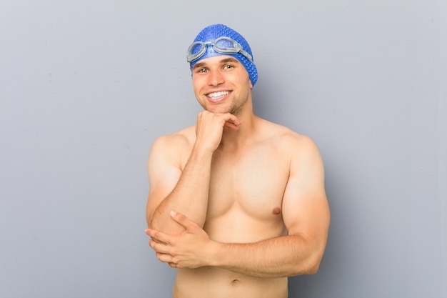 Hombre joven nadador profesional sonriendo feliz y confiado, tocando la barbilla con la mano.