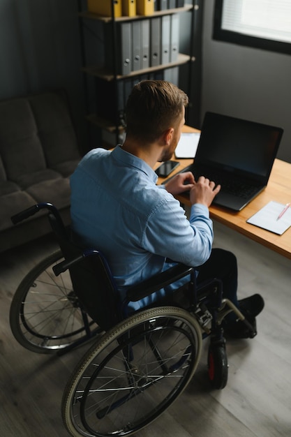 Hombre joven con necesidades especiales en ropa informal que trabaja en una computadora portátil inalámbrica Hombre independiente que trabaja desde casa mientras está sentado en silla de ruedas