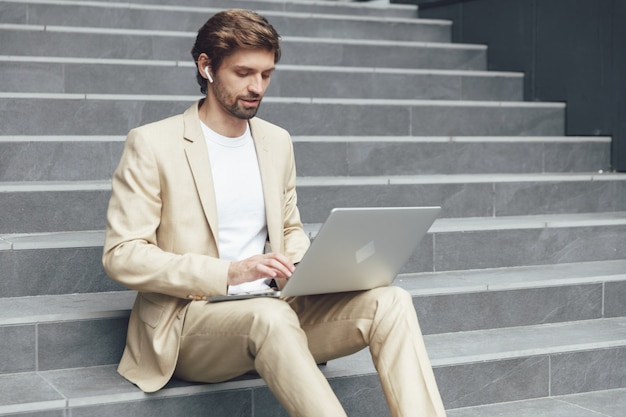 Hombre joven ocupado en traje de moda escribiendo en la computadora portátil moderna mientras está sentado en los pasos afuera