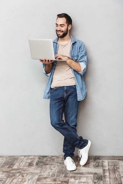 Hombre joven optimista de longitud completa con barba escribiendo en la computadora portátil y sonriendo aislado sobre pared gris