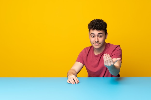 Hombre joven con pared colorida y mesa invitando a venir con la mano. Feliz de que hayas venido