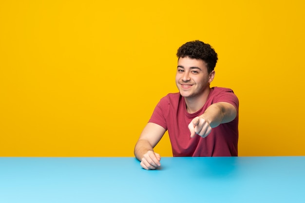 Hombre joven con pared colorida y mesa te señala con una expresión segura