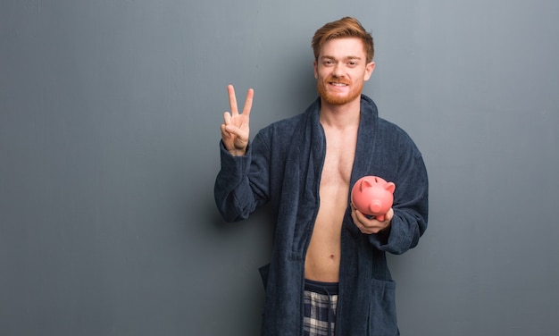 Hombre joven pelirrojo en pijama mostrando el número dos. Tiene una alcancía.