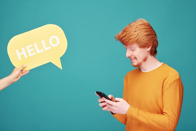 Hombre joven pelirrojo positivo mirando a un lado inquisitivamente y sosteniendo pancarta de tweet contra la pared azul