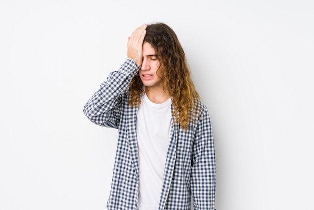 Hombre joven de pelo largo posando aislado olvidando algo, golpeándose la frente con la palma y cerrando los ojos.