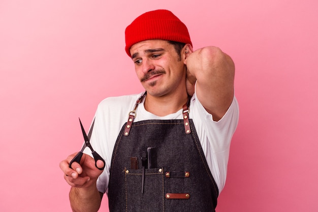 Hombre joven del peluquero caucásico que sostiene las tijeras aisladas en fondo rosado que toca la parte posterior de la cabeza, pensando y haciendo una elección.