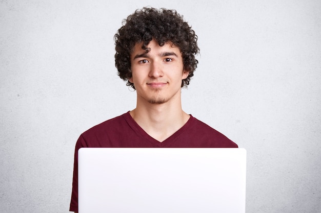 Hombre joven pensativo con cabello rizado, mira directamente a la cámara, usa una computadora portátil blanca moderna, viste una camiseta granate informal, se sienta contra la pared de concreto blanco con espacio en blanco para publicidad.