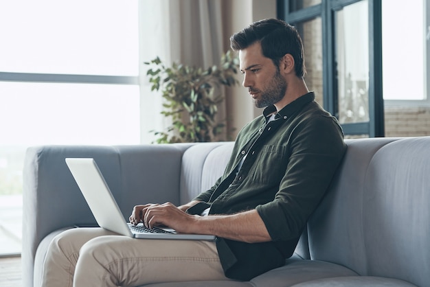 Hombre joven pensativo usando la computadora portátil mientras está sentado en el sofá