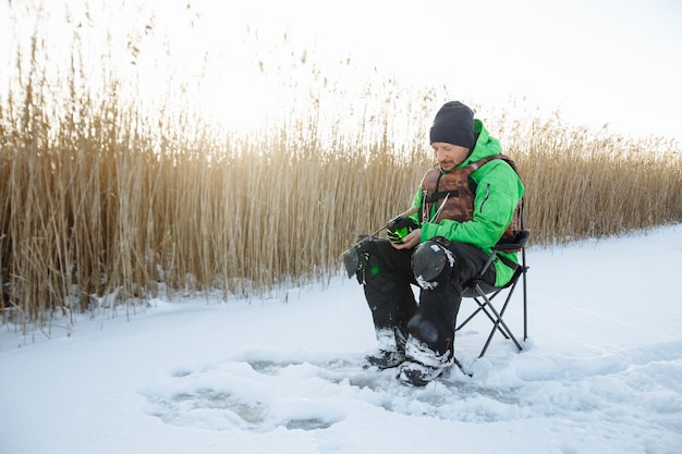 Un hombre joven en un pescador de invierno se sienta en una silla y pesca y una caña de pescar