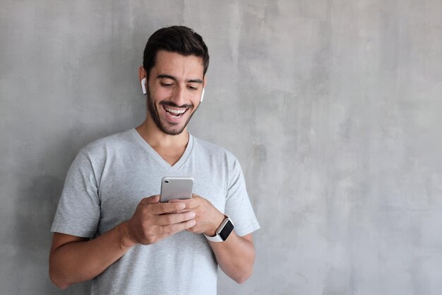 Foto hombre joven de pie contra la pared de textura gris con espacio de copia para anuncios mirando agitada en la exhibición de su teléfono inteligente impresionado por el contenido de los medios