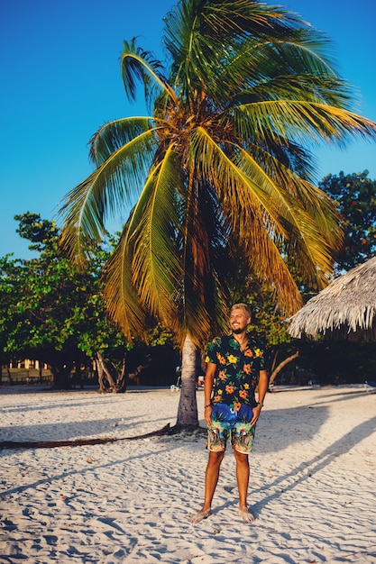 Hombre joven de pie en la playa mientras sonríe contra el fondo de las palmeras
