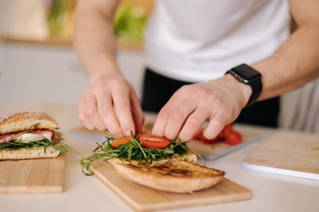 Hombre joven pone tomates en sándwich Hombre preparando el almuerzo en casa