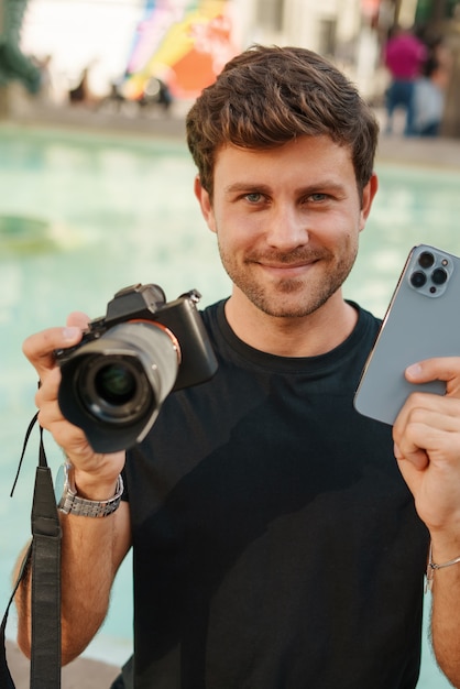 Hombre joven positivo que sostiene el teléfono inteligente y la cámara de fotos en las manos y sonriendo
