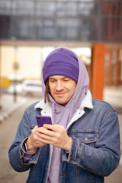 Hombre joven positivo en ropa de abrigo sonriendo y navegando por teléfono móvil mientras está de pie en la calle de la ciudad
