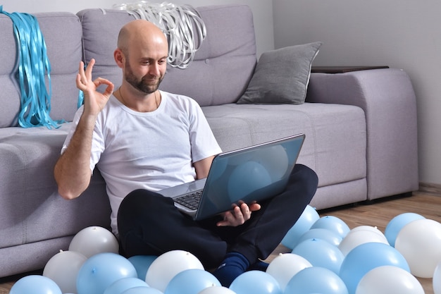 Hombre joven positivo en ropa casual sentado en la sala de estar y usando la computadora portátil