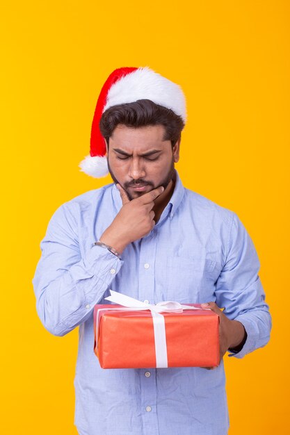 Hombre joven positivo Santa Claus con regalos y árbol de Navidad posando sobre una pared amarilla. Concepto de vacaciones de Navidad y año nuevo.