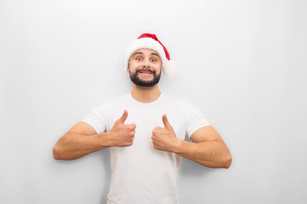 Hombre joven positivo se ve. Él muestra sus grandes pulgares hacia arriba. Guy usa sombrero de Chritmas.