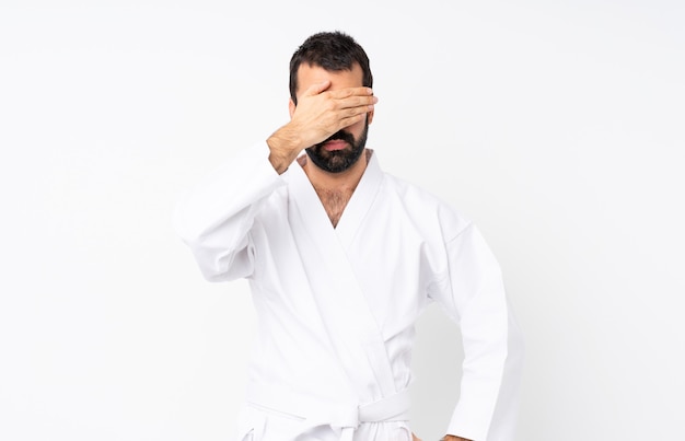 Hombre joven que hace karate sobre blanco aislado que cubre los ojos con las manos. No quiero ver algo