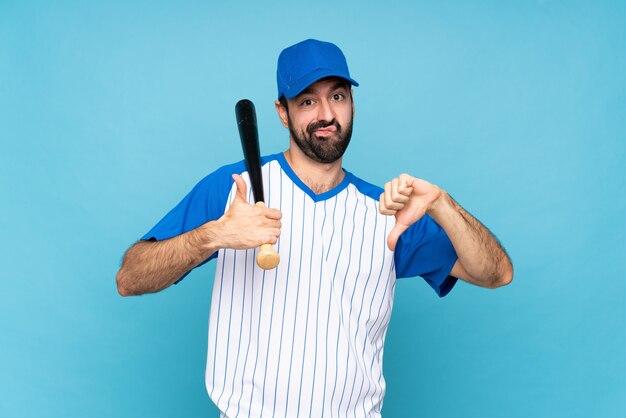 Hombre joven que juega a béisbol sobre azul aislado que hace la muestra buena-mala. Indeciso entre sí o no