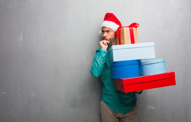 Hombre joven que lleva un sombrero de santa que sostiene los regalos que piensa en una idea