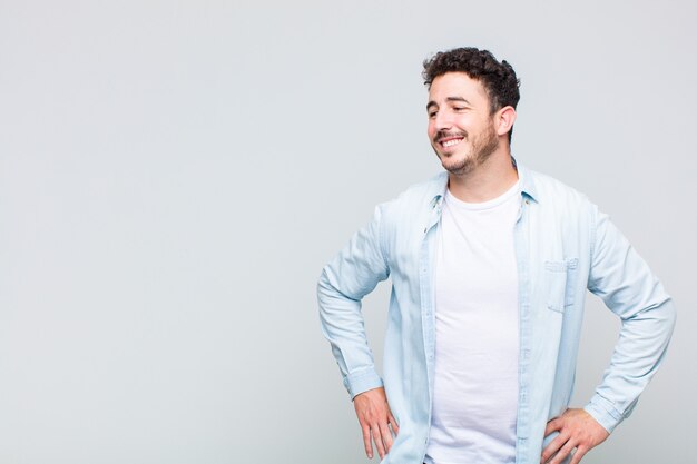 Hombre joven que parece feliz, alegre y confiado, sonriendo con orgullo y mirando al lado con ambas manos en las caderas