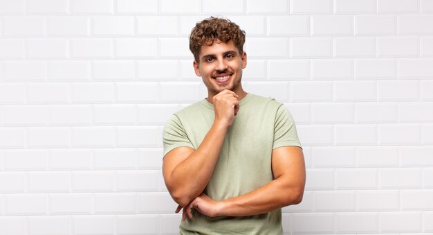 Hombre joven que parece feliz y sonriente con la mano en la barbilla, preguntándose o haciendo una pregunta, comparando opciones