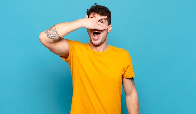 Hombre joven que parece sorprendido, asustado o aterrorizado, cubriendo la cara con la mano y mirando entre los dedos