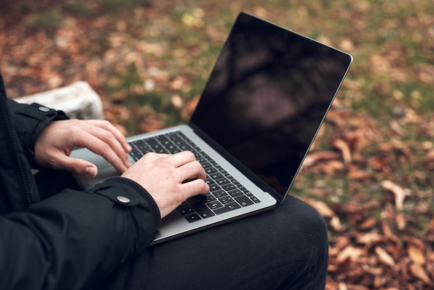 Hombre joven que se sienta en banco de parque en otoño con la computadora portátil. Estudiante usando la computadora al aire libre. De cerca.