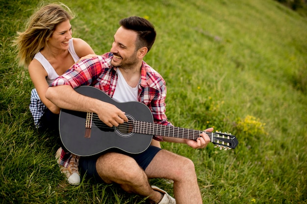 Hombre joven que se sienta en hierba con su novia y que toca la guitarra