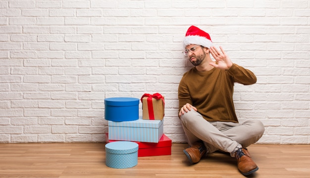 Hombre joven que se sienta con los regalos que celebran la Navidad que pone la mano en frente