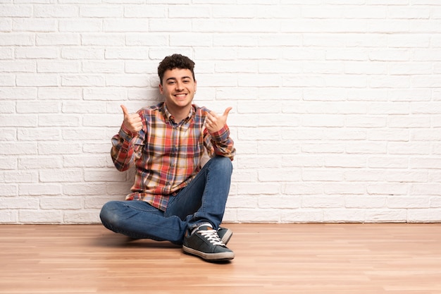 Hombre joven que se sienta en el suelo con los pulgares para arriba gesto y la sonrisa