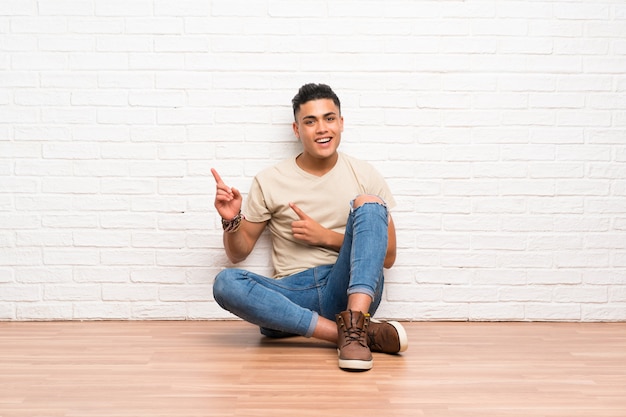 Hombre joven que se sienta en el suelo que señala el dedo al lado