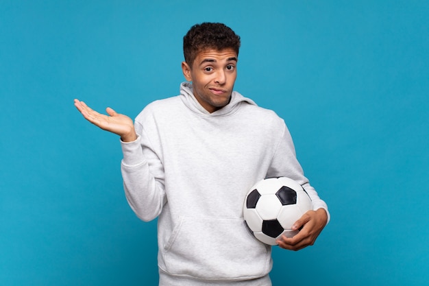 Hombre joven que se siente desconcertado y confundido, dudando, ponderando o eligiendo diferentes opciones con expresión divertida. concepto de futbol