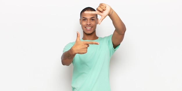 Hombre joven que se siente feliz, amigable y positivo, sonriendo y haciendo un retrato o un marco de fotos con las manos