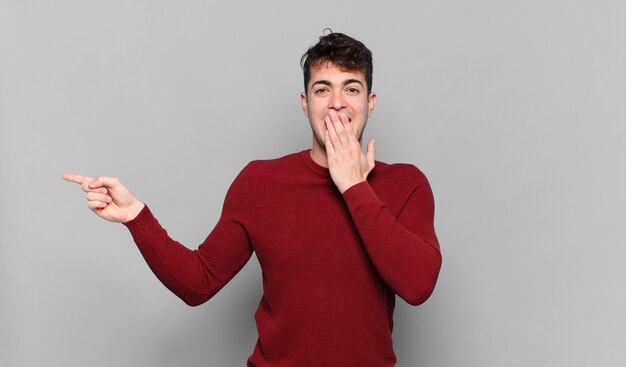 Hombre joven que se siente feliz, conmocionado y sorprendido, cubriendo la boca con la mano y apuntando al espacio de copia lateral