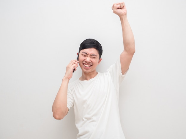 Hombre joven que se siente feliz y exitoso sosteniendo el teléfono celular y la mano hacia arriba el fondo de la pared blanca