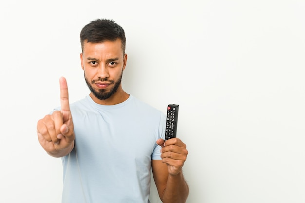 Hombre joven que sostiene un controlador de televisión que muestra el número uno con el dedo.