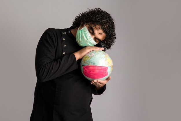 Hombre joven que sostiene el globo del mundo con una mascarilla de la medicina. Epidemia mundial del concepto de coronavirus.