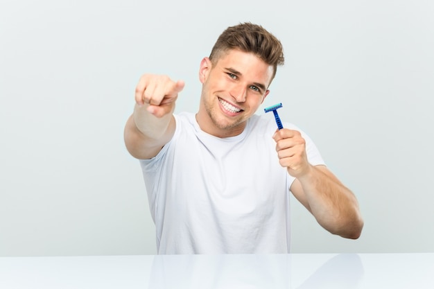 Foto hombre joven que sostiene una hoja de afeitar alegres sonrisas apuntando al frente.
