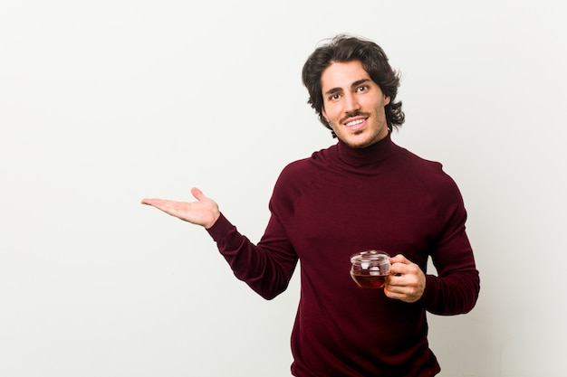 Hombre joven que sostiene una taza de té que muestra un espacio en blanco en una palma y que sostiene otra mano en la cintura.