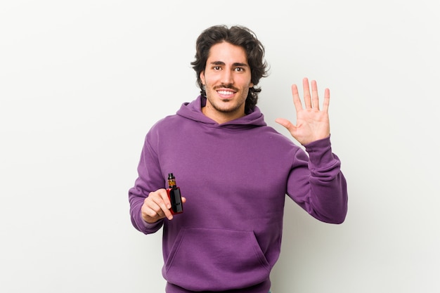 Hombre joven que sostiene un vaporizador sonriente alegre que muestra el número cinco con los dedos.