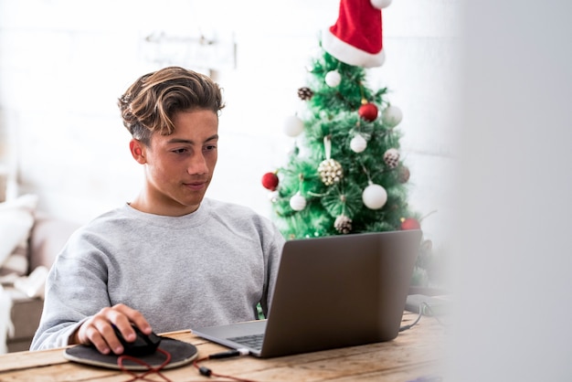 Un hombre joven que usa su computadora portátil y computadora en casa sentado en una silla y trabajando en la mesa solo con el árbol de Navidad en el fondo - adolescente feliz disfrutando de vacaciones y vacaciones