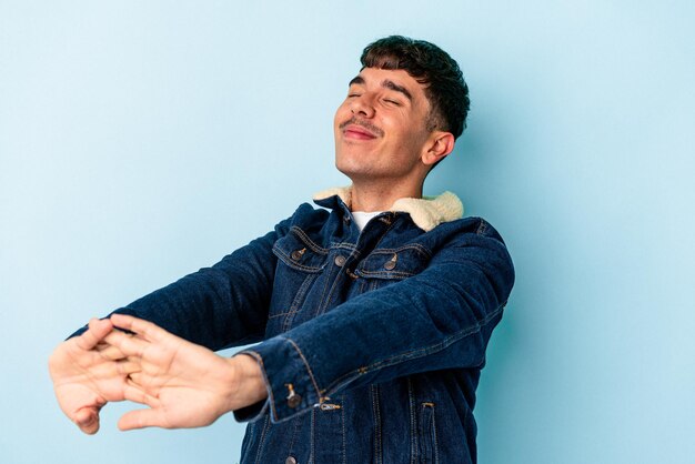 Hombre joven de raza mixta aislado sobre fondo azul estirando los brazos, posición relajada.