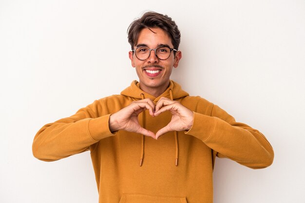 Hombre joven de raza mixta aislado sobre fondo blanco sonriendo y mostrando una forma de corazón con las manos.