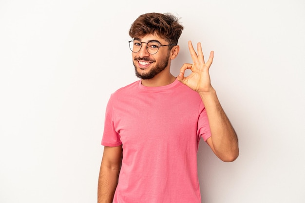 Hombre joven de raza mixta aislado sobre fondo gris alegre y confiado mostrando gesto ok.