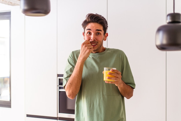 Hombre joven de raza mixta bebiendo jugo de naranja en su cocina mordiéndose las uñas, nervioso y muy ansioso.