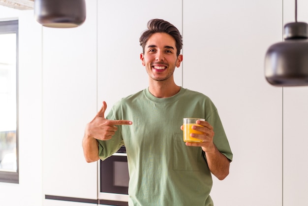 Hombre joven de raza mixta bebiendo jugo de naranja en su cocina persona apuntando con la mano a un espacio de copia de camisa, orgulloso y seguro